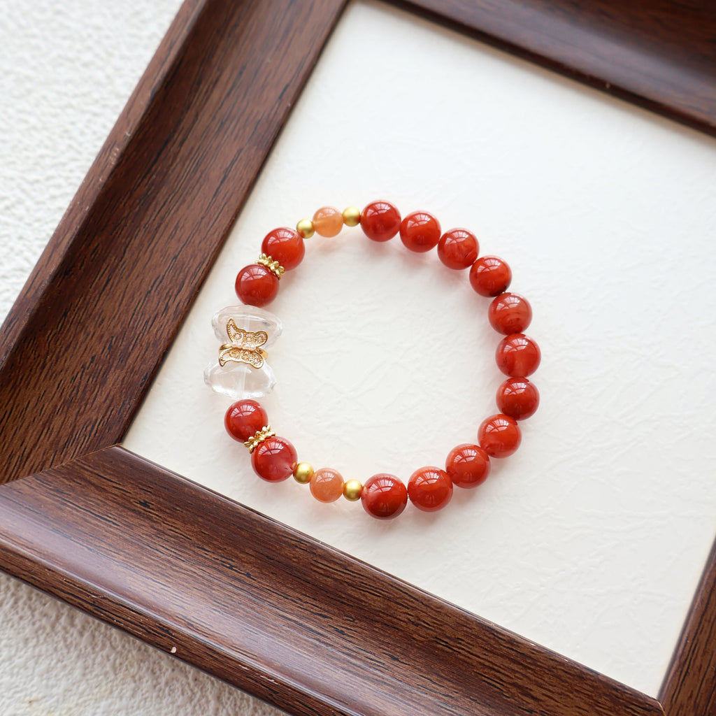 Beaded bracelet with red beads and gold accents on a white surface, framed by a wooden border.