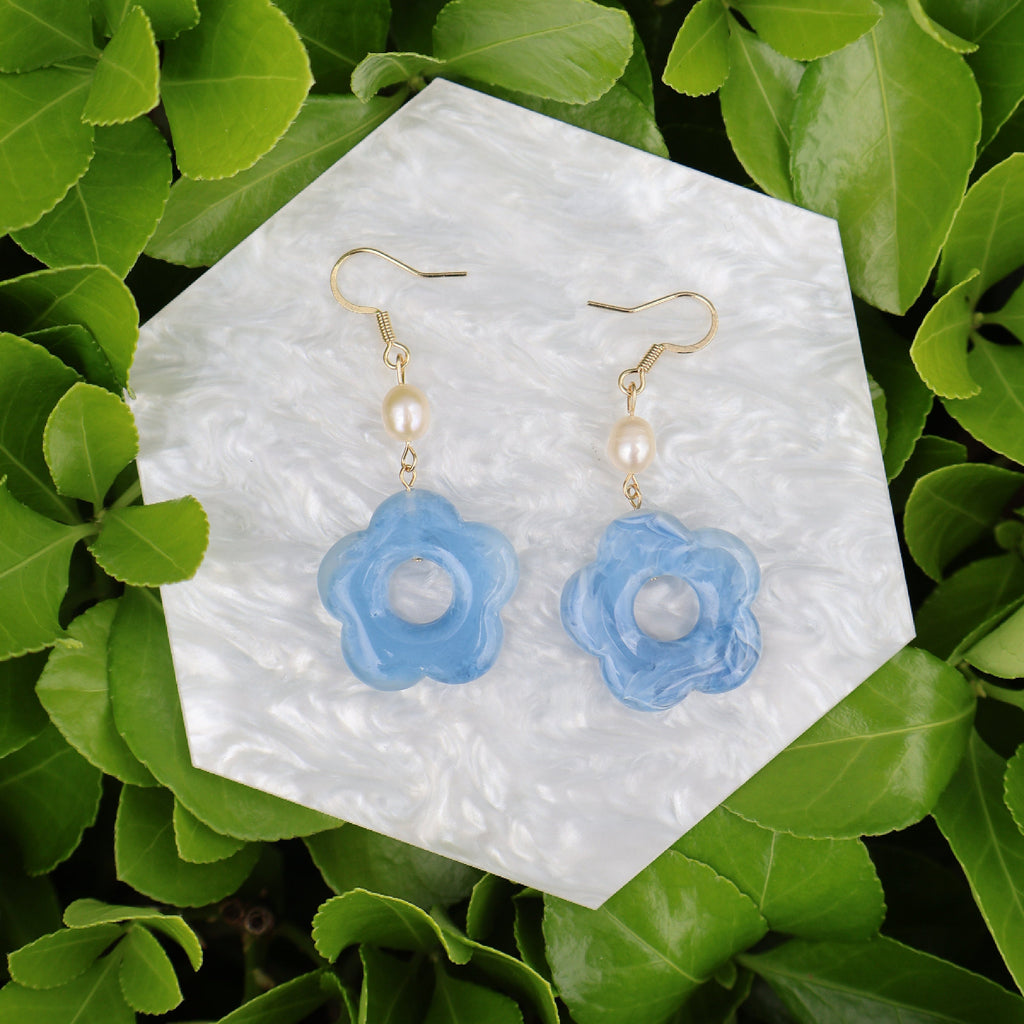 Blue flower-shaped earrings on a white hexagonal stone with green leaves in the background