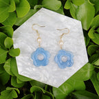 Blue flower-shaped earrings on a white hexagonal stone with green leaves in the background