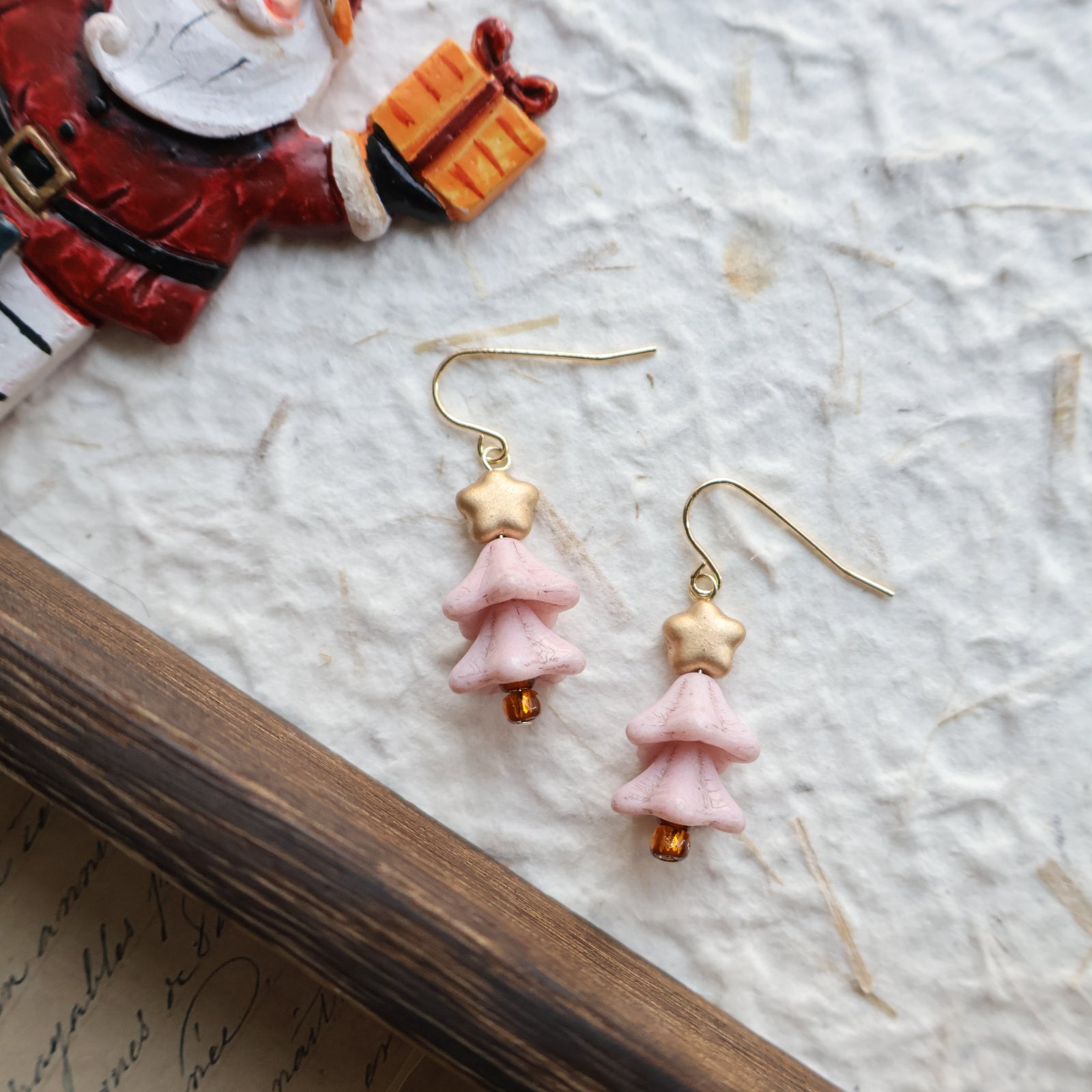 Pair of pink and gold earrings on a textured surface with a book and decorative item in the background.