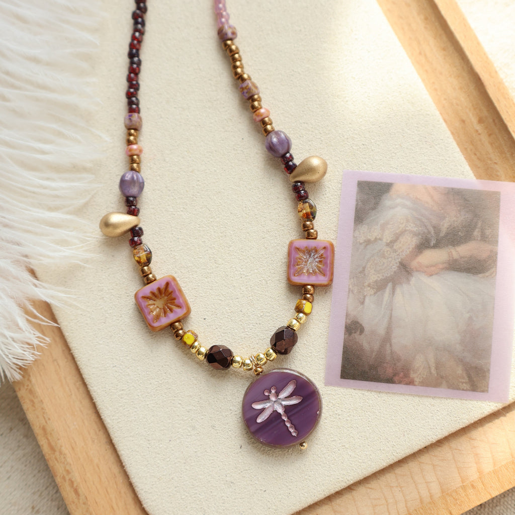 Necklace with colorful beads and a dragonfly charm on a wooden tray with a photo card.