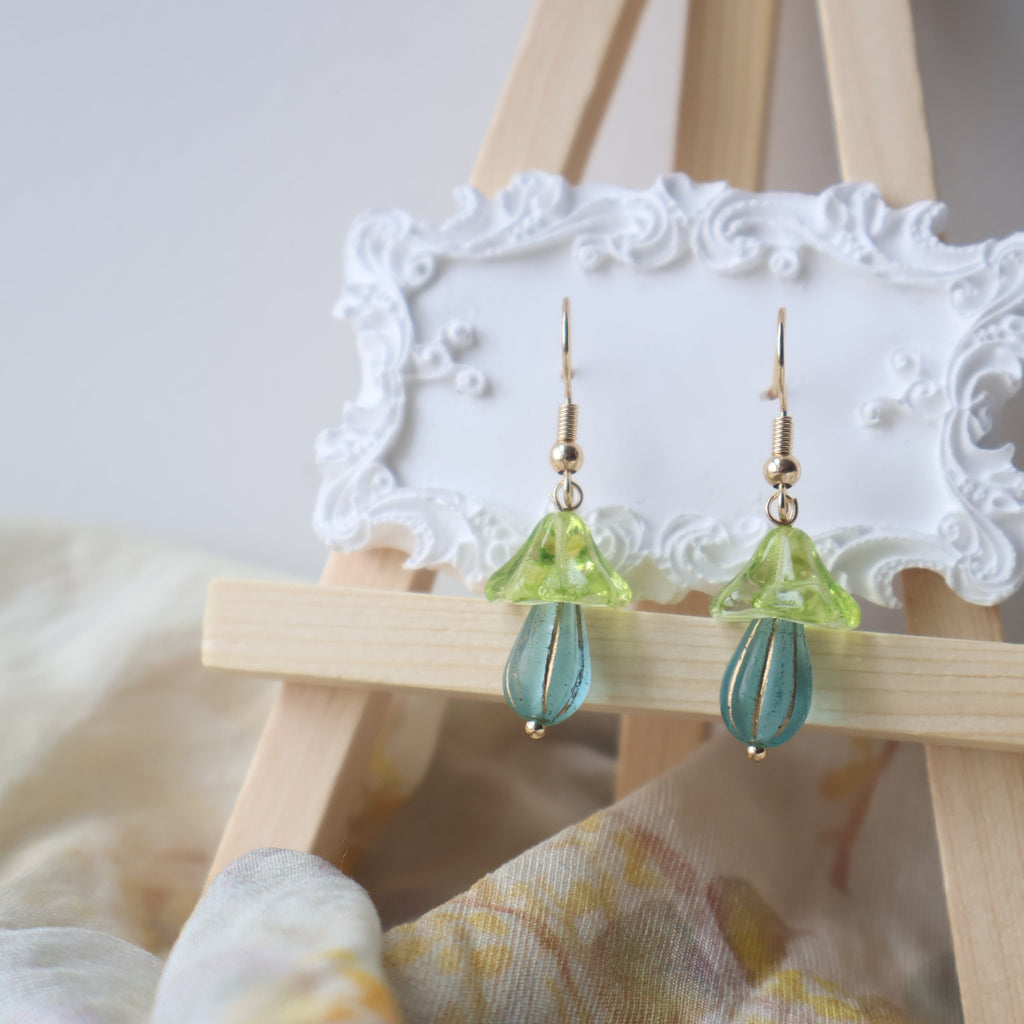 Pair of green and blue beaded earrings on a wooden stand with a white lace background.