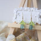 Pair of green and blue beaded earrings on a wooden stand with a white lace background.