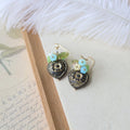 Pair of decorative earrings on a textured surface with a feather and paper in the background