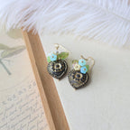 Pair of decorative earrings on a textured surface with a feather and paper in the background