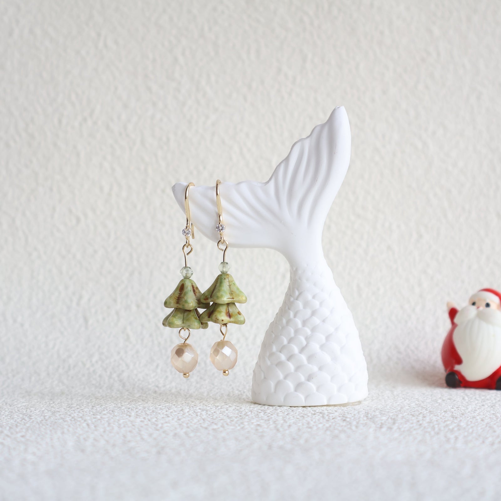 Earrings with green and beige elements on a white mermaid tail stand against a light background.