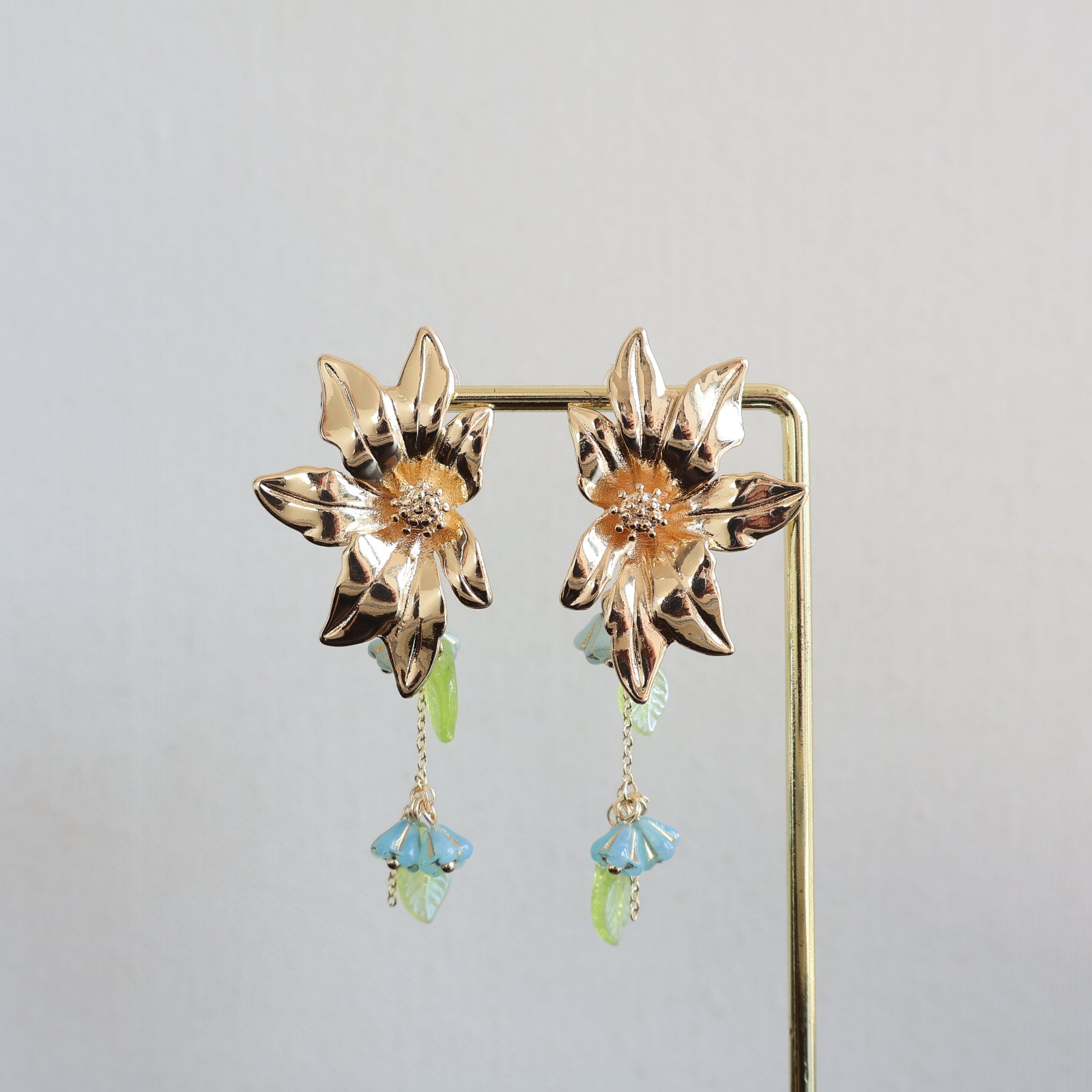 Gold flower-shaped earrings with green and blue drops on a gray background
