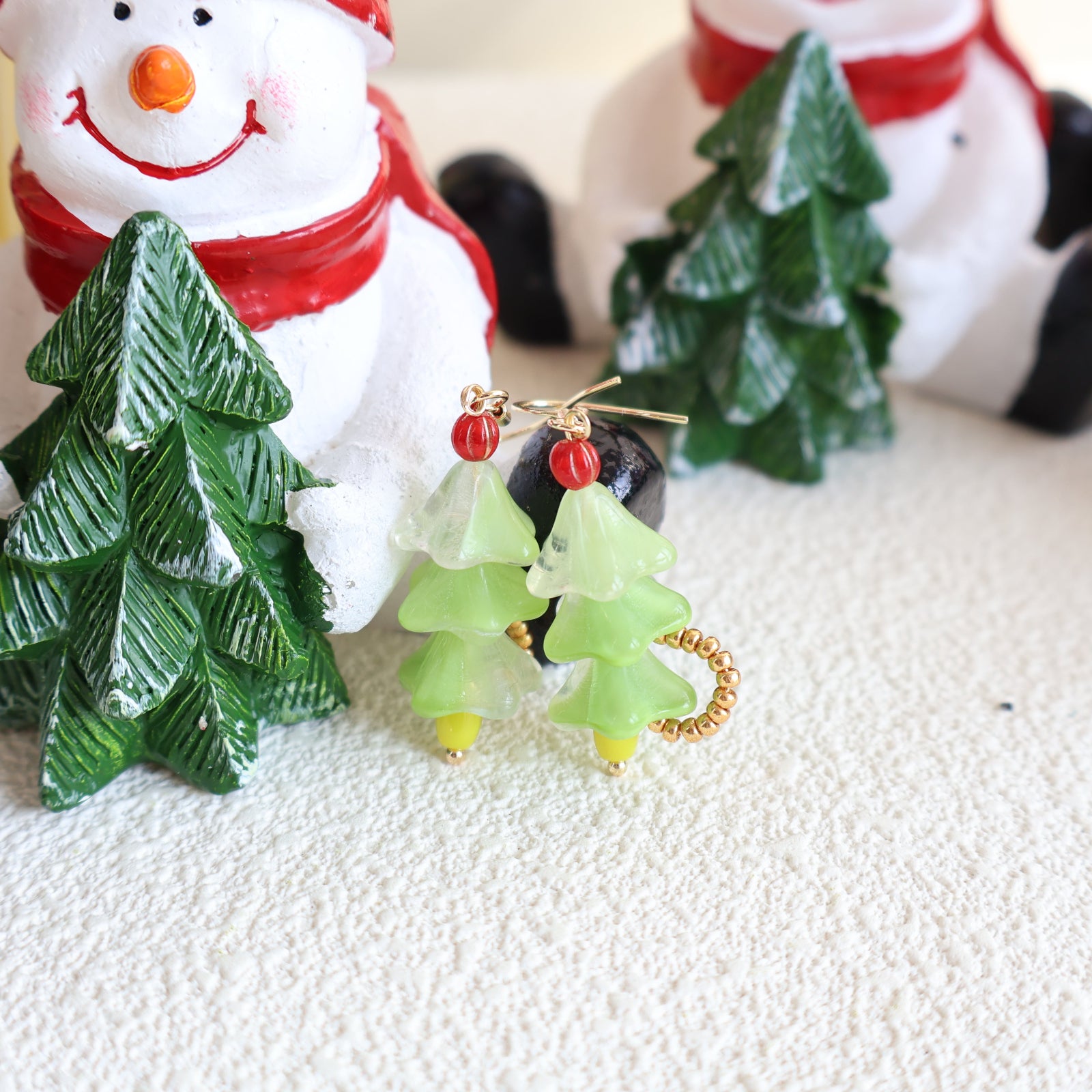 Green Christmas tree-shaped earrings with red berries in front of a snowman and small trees decoration.