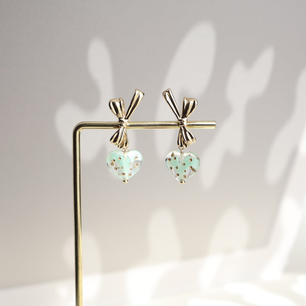 Pair of earrings with heart-shaped green charms on a stand against a neutral background
