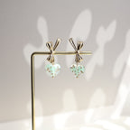 Pair of earrings with heart-shaped green charms on a stand against a neutral background
