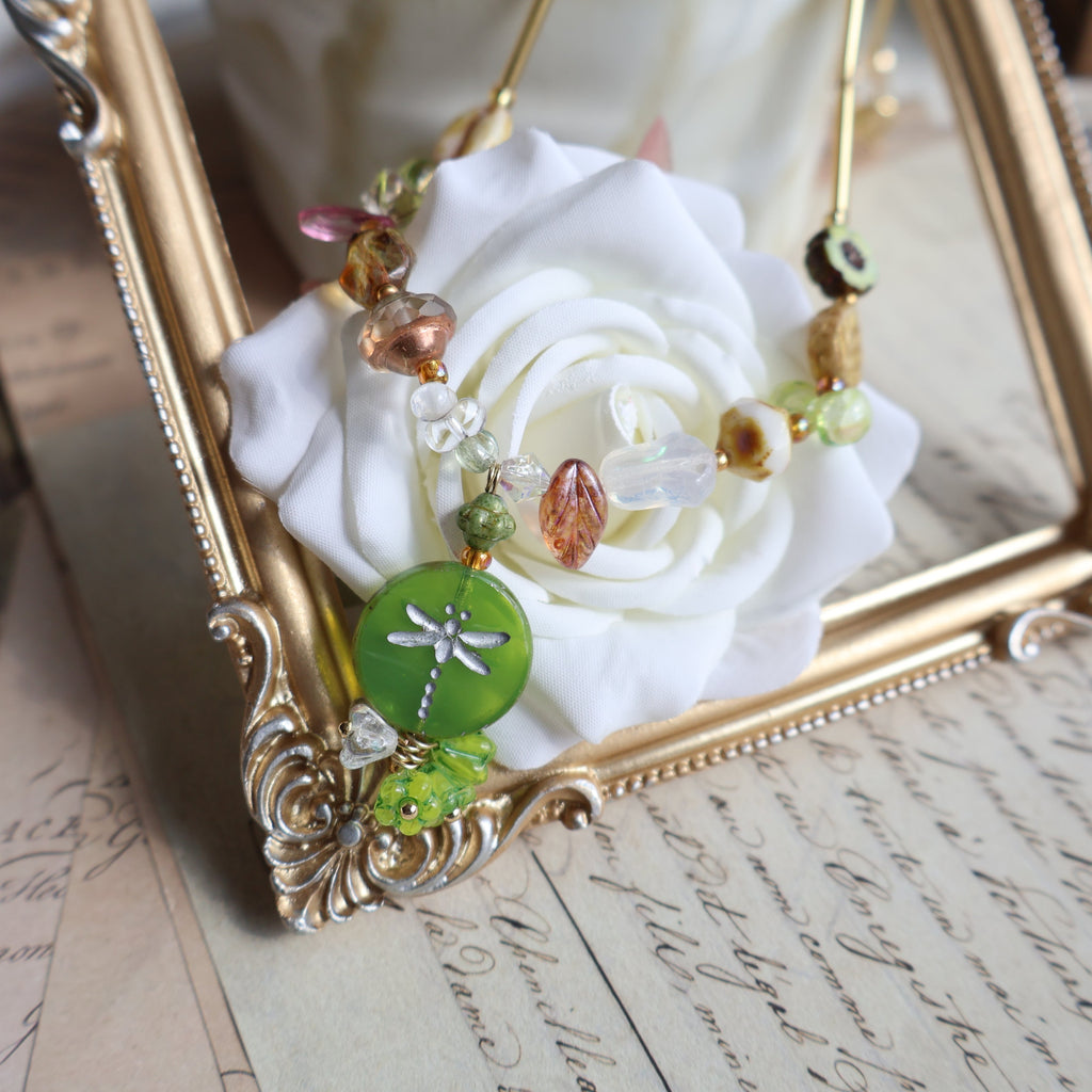 Decorative bracelet with beads and a green charm on a white rose, set against an ornate gold frame.