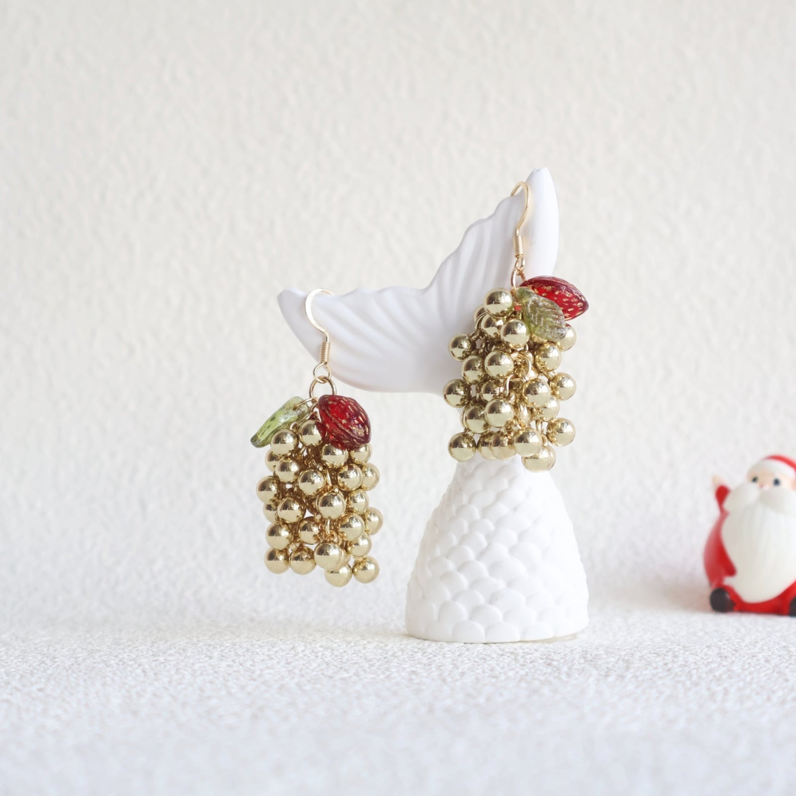 Decorative earrings with fruit and gold bead details on a white stand with a Santa Claus figure in the background.