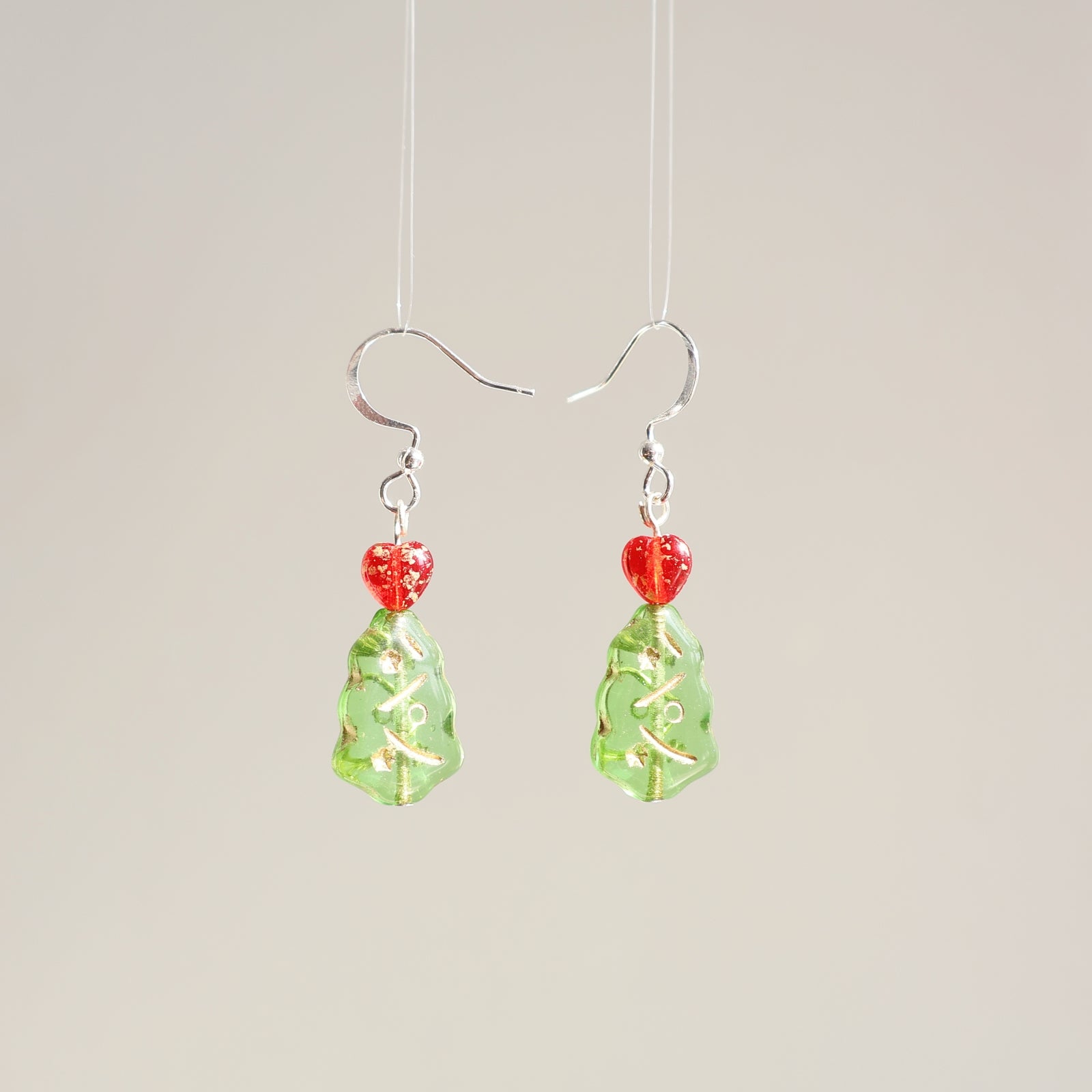Pair of earrings with green teardrop-shaped beads and red heart-shaped accents on a beige background.