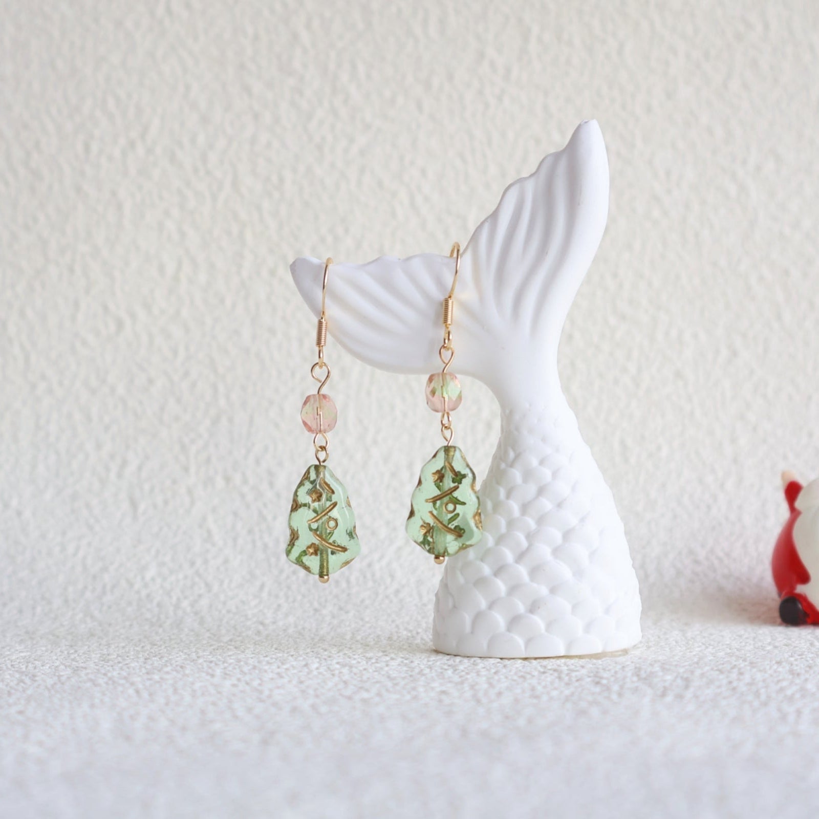 Pair of earrings hanging from a white mermaid tail stand on a textured white surface.
