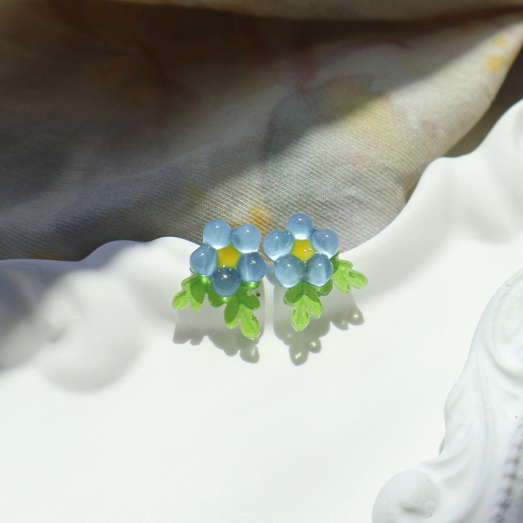 Blue flower-shaped earrings on a white surface with a beige fabric background