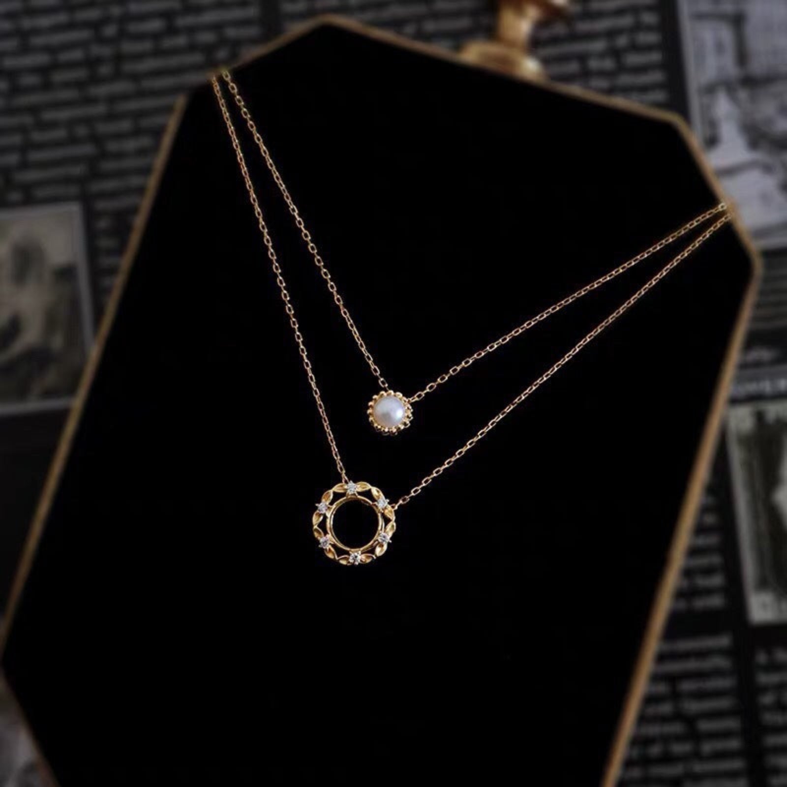 Two gold necklaces on a black stand with a blurred vintage-style background