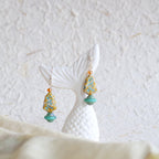 Pair of earrings with turquoise and gold beads on a white stand against a textured white background