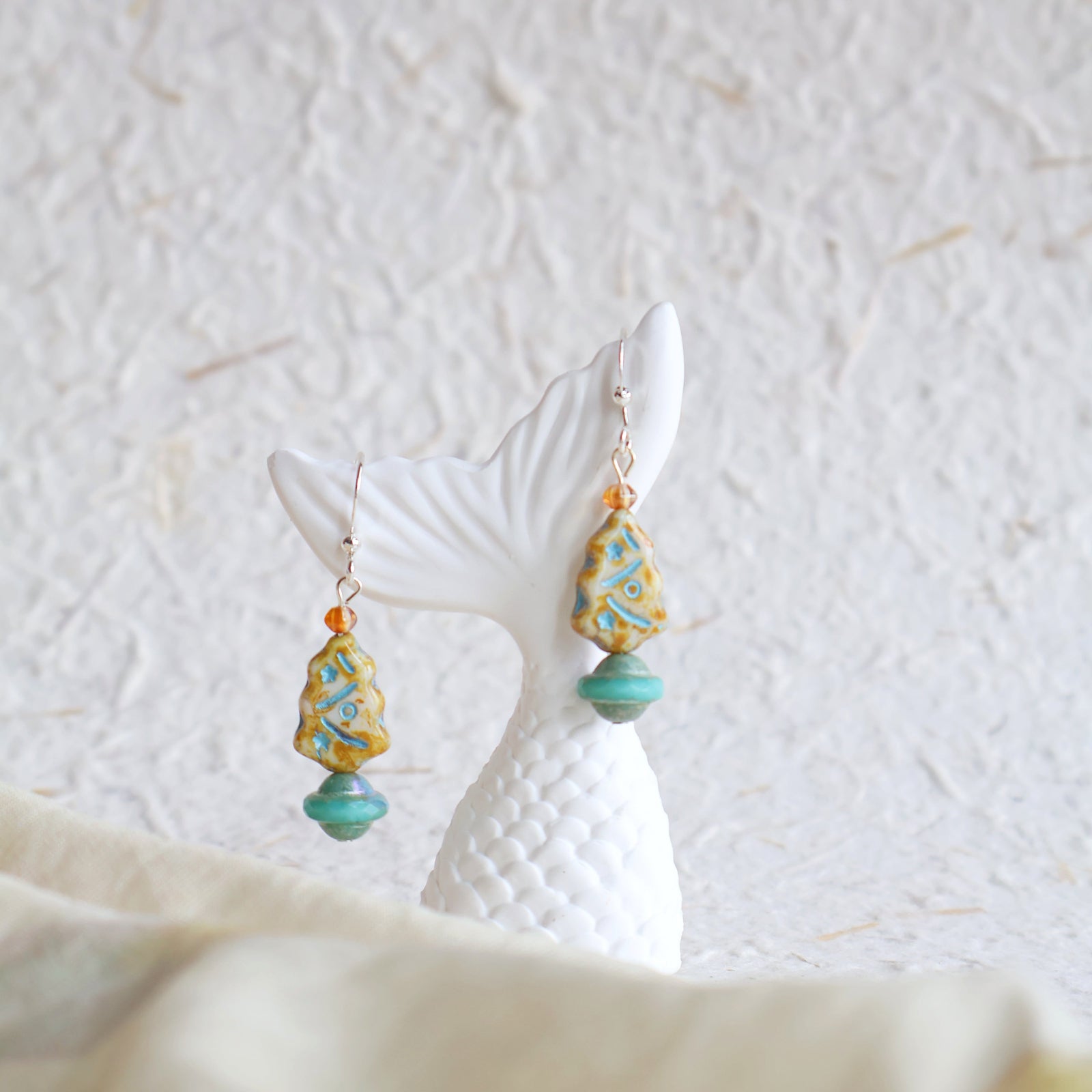 Pair of earrings with turquoise and gold beads on a white stand against a textured white background