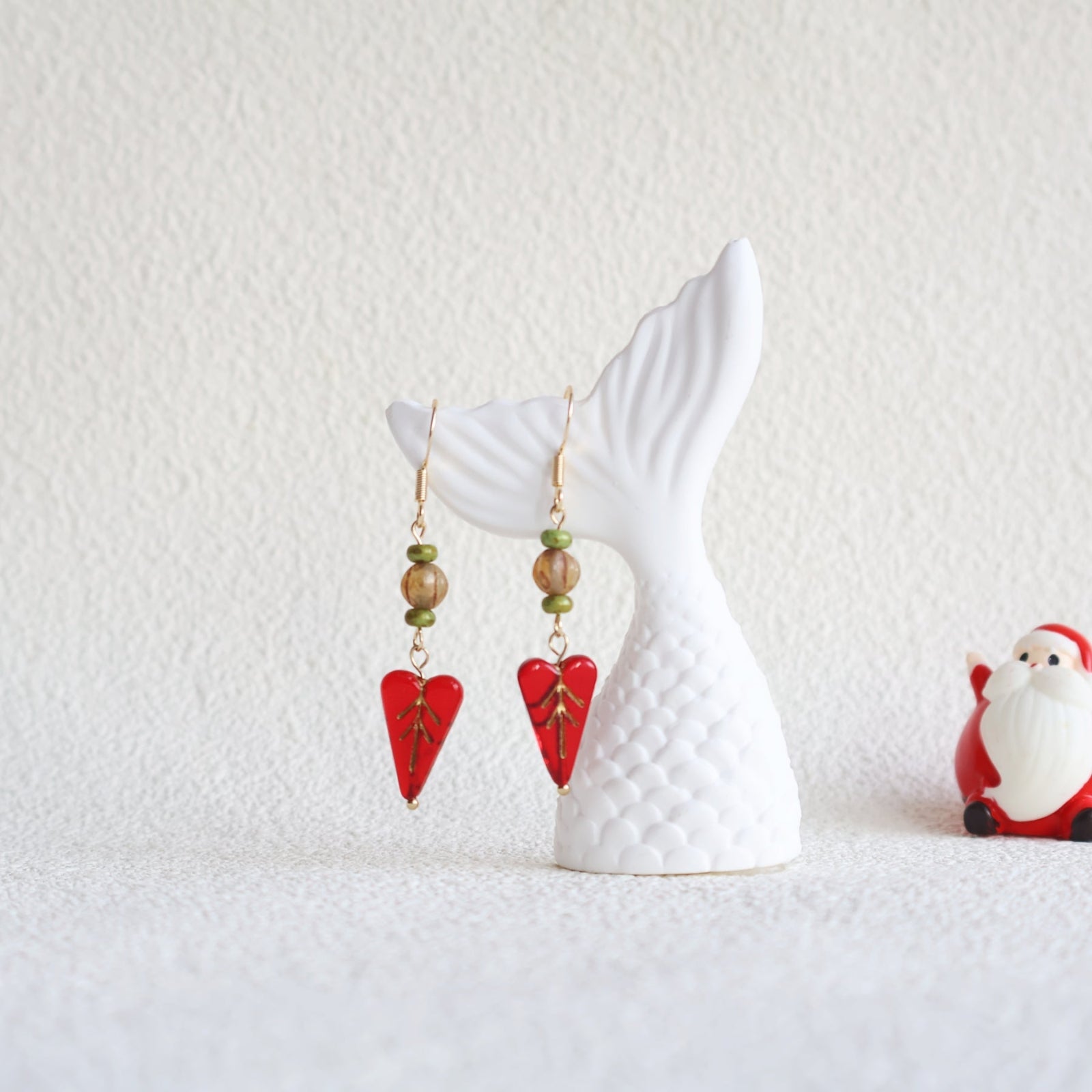 Red heart-shaped earrings with green beads on a white horse-shaped stand against a white background.